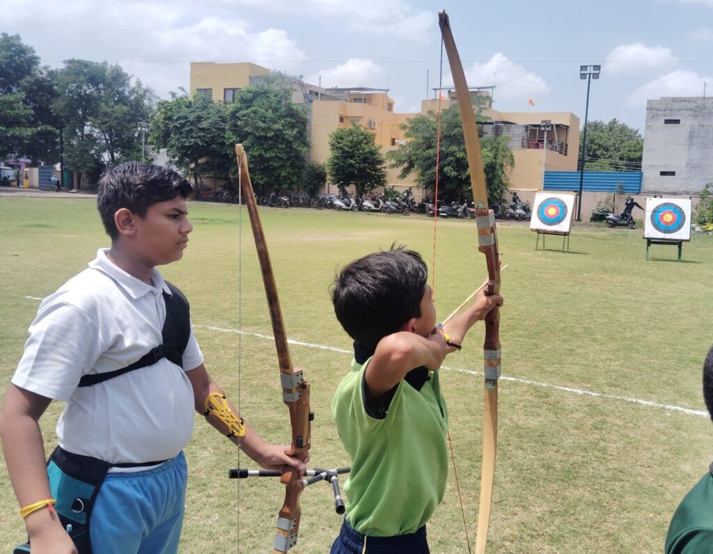 तीरंदाजी प्रतियोगिता में छात्र तीर चलाते हुए; शिक्षकों और अधिकारियों की उपस्थिति में कार्यक्रम का शुभारंभ